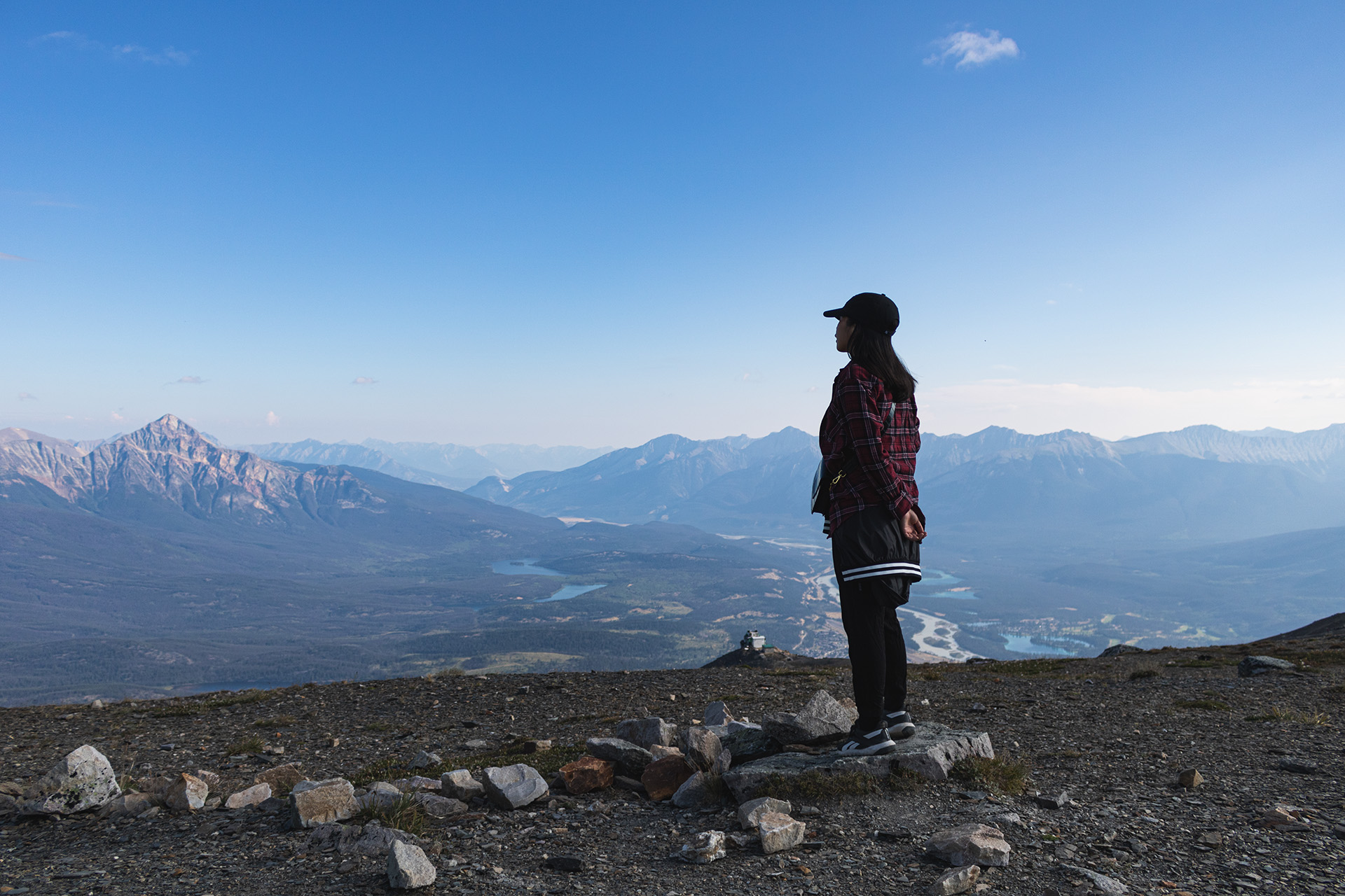 Girl standing on a mountain plateau and looking towards a large valley with small lakes and rocky mountains in the distance.